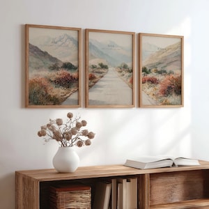 May include: Three framed landscape prints with text: "Ask", "Seek", and "Knock". Each print features a road winding through a mountain scene with muted colors. The frames are a light brown wood. A white vase with dried flowers sits on a wooden shelf.