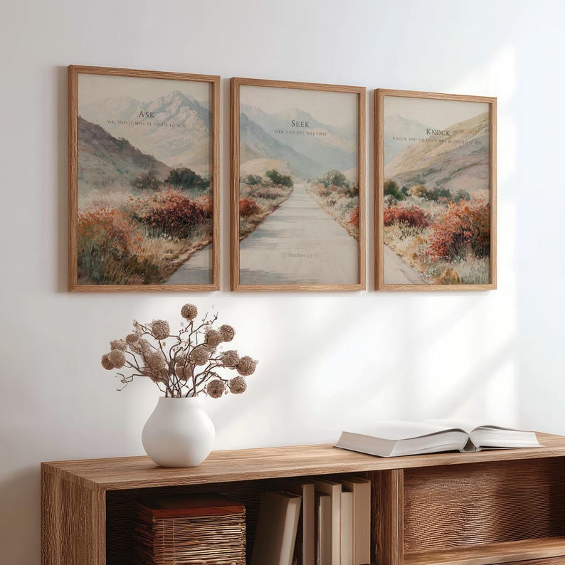 May include: Three framed landscape prints with text: "Ask", "Seek", and "Knock". Each print features a road winding through a mountain scene with muted colors. The frames are a light brown wood. A white vase with dried flowers sits on a wooden shelf.