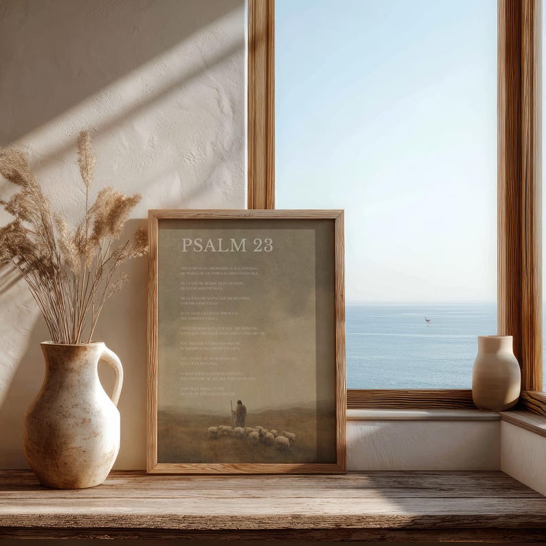 May include: A framed print with the text "PSALM 23" in a wooden frame. The print depicts a shepherd with sheep in a field. A vase with dried plants and a small vase are on a wooden surface near a window with a view of the ocean.