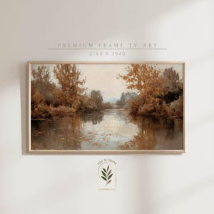 Puede incluir: Una pintura de paisaje enmarcada que presenta una serena escena de río con árboles en colores otoñales. La obra de arte tiene una estética vintage, con texto en la parte inferior que dice "He leads me beside still waters, He restores my soul."