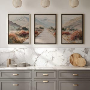 May include: Three framed landscape art prints with inspirational text. Each print features a mountain scene with a road or path. The text on the prints reads "Ask", "Seek", and "Knock". The frames are gold, and the prints are displayed above a gray cabinet.
