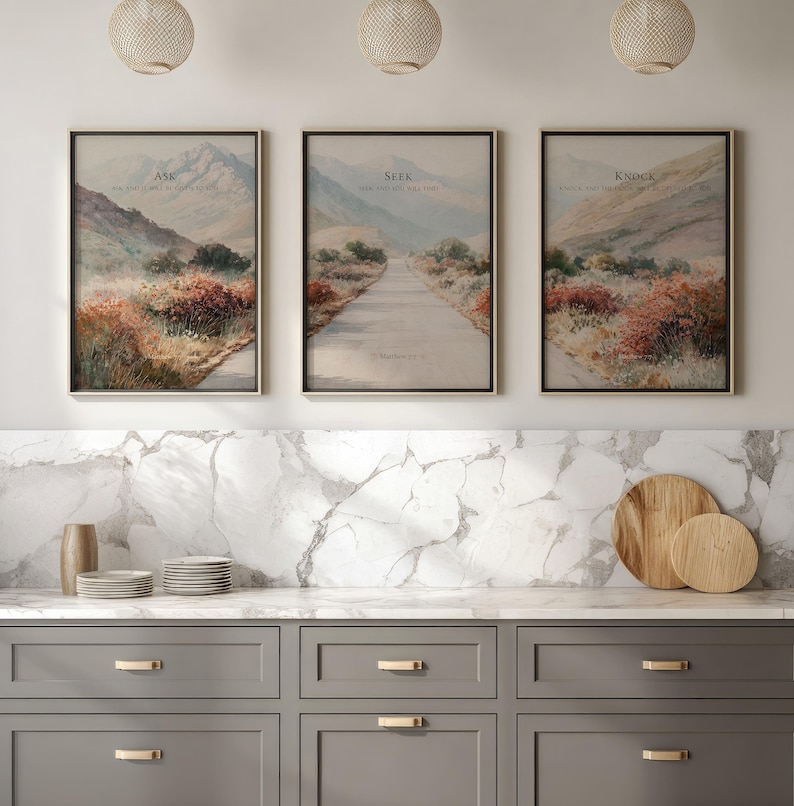 May include: Three framed landscape art prints with inspirational text. Each print features a mountain scene with a road or path. The text on the prints reads "Ask", "Seek", and "Knock". The frames are gold, and the prints are displayed above a gray cabinet.