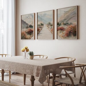 May include: Three framed landscape prints with text, "Ask", "Seek", and "Knock", hang above a dining table. The prints depict a road through a mountain landscape with muted colors. A vase of yellow flowers and a potted plant sit on the table.