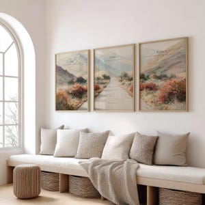 May include: Three framed landscape art prints with the words "Ask", "Seek", and "Knock" in a light beige frame. The prints depict a road winding through a mountain landscape with muted colors. The room also features a bench with pillows and a woven basket.