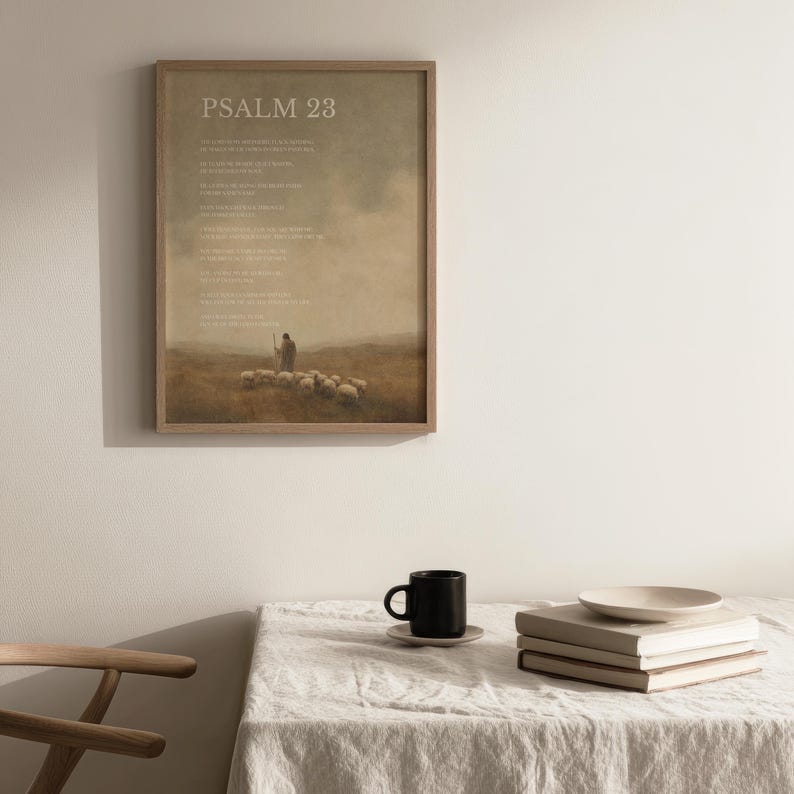 May include: A framed print of Psalm 23, featuring a pastoral scene with a shepherd and sheep. The artwork has a muted color palette with a beige background and a wooden frame. A black mug, saucer, books, and a plate are on a table.