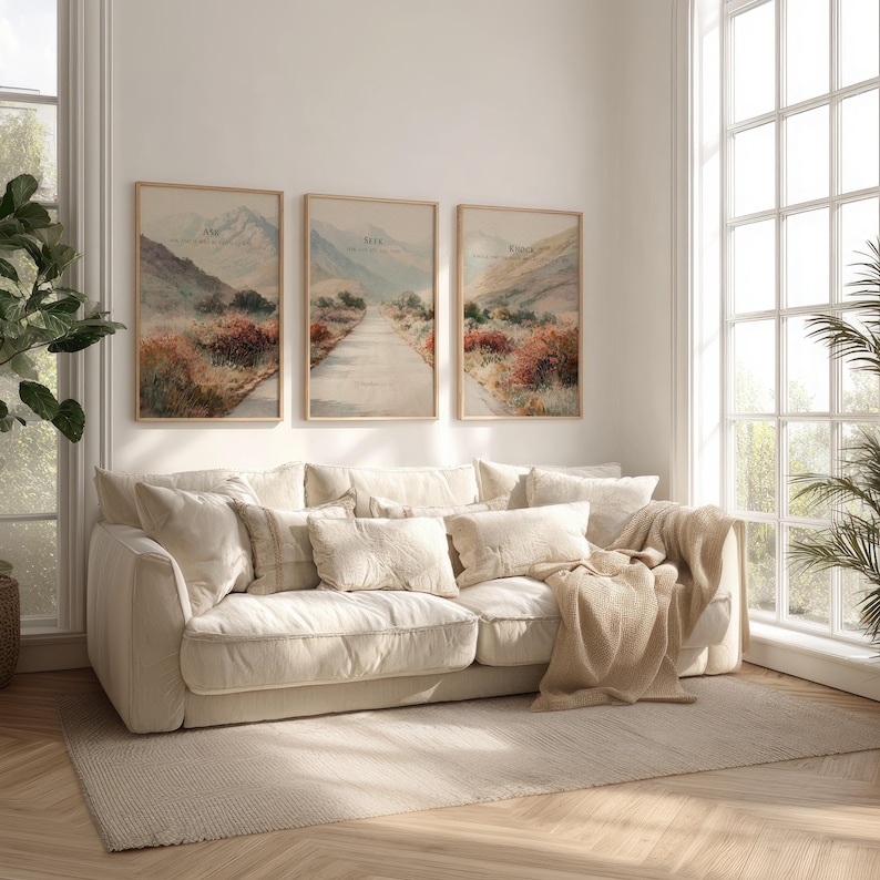 May include: Three framed landscape art prints with a neutral color palette hang above a cream-colored sofa. The prints depict a road winding through a mountain scene with text at the top. The sofa is adorned with multiple pillows and a beige throw blanket.