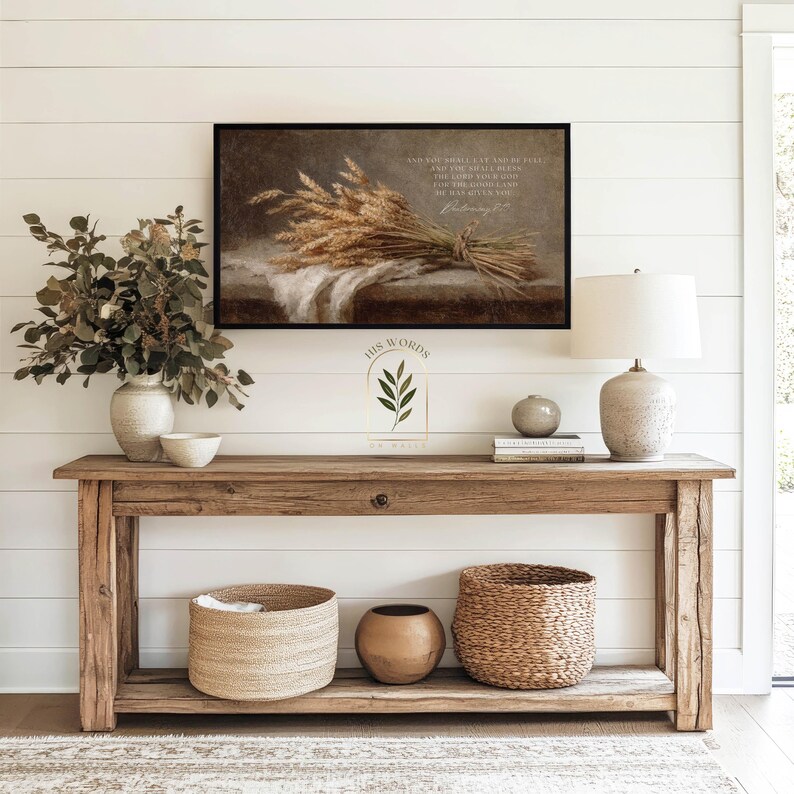 May include: A rustic wooden console table with decorative items. A framed print with a wheat bundle and text hangs above. A vase with greenery, a lamp, and woven baskets complete the decor. The text on the print reads: "AND YOU SHALL LAY UP..."