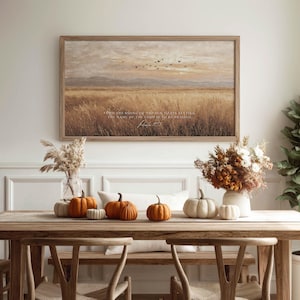 May include: A framed landscape print with a field of tall grass and a sunset sky. The print is above a wooden table decorated with pumpkins and floral arrangements. The text on the print reads, "From the rising of the sun..."