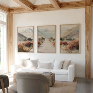 May include: Three framed landscape art prints with text that reads "Ask", "Seek", and "Knock". The prints feature a road, mountains, and colorful foliage. The artwork is displayed above a white sofa in a living room.