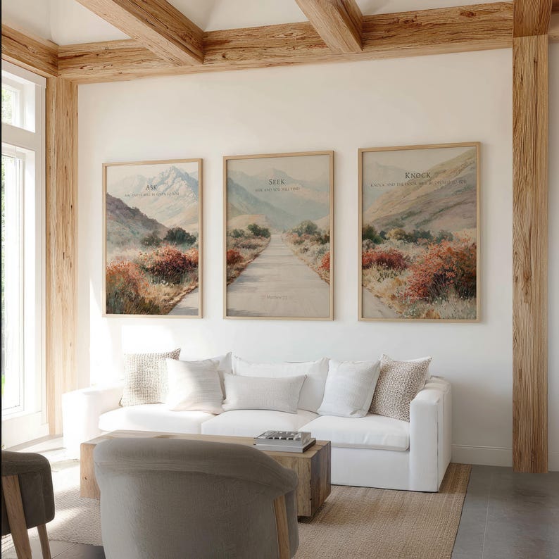 May include: Three framed landscape art prints with text that reads "Ask", "Seek", and "Knock". The prints feature a road, mountains, and colorful foliage. The artwork is displayed above a white sofa in a living room.
