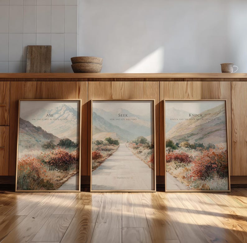 May include: Three framed landscape art prints with inspirational text. Each print features a watercolor painting of a path leading through a mountain valley. The text on the prints reads "Ask", "Seek", and "Knock". The frames are a light brown color.