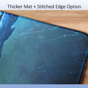May include: A rectangular desk mat featuring a dark blue and teal dragon graphic with a glowing red eye. The mat has a stitched edge and the text Thicker Mat + Stitched Edge Option is displayed above the product in the image frame.