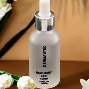 Puede incluir: Una botella de vidrio esmerilado con un cuentagotas plateado, etiquetada como "DERMABTEC HYALURONIC ACID SERUM SKIN CARE SERUM". La botella está rodeada de flores blancas y una margarita sobre un fondo marrón y beige.