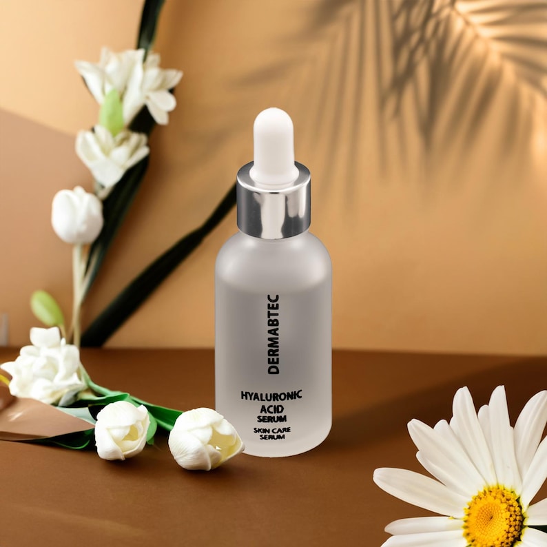 May include: A frosted glass bottle with a silver dropper top, labeled "DERMABTEC HYALURONIC ACID SERUM SKIN CARE SERUM." The bottle is surrounded by white flowers and a daisy against a brown and tan background.