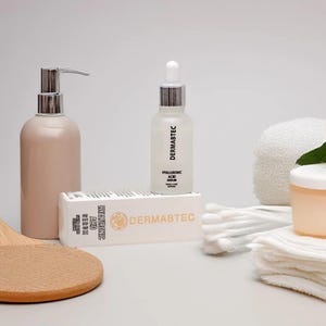 May include: A skincare product arrangement featuring a light pink pump bottle, a clear serum bottle with a dropper, and a box labeled "DERMABTEC HYALURONIC ACID SERUM". Additional items include cotton swabs, a small jar, and a white towel.