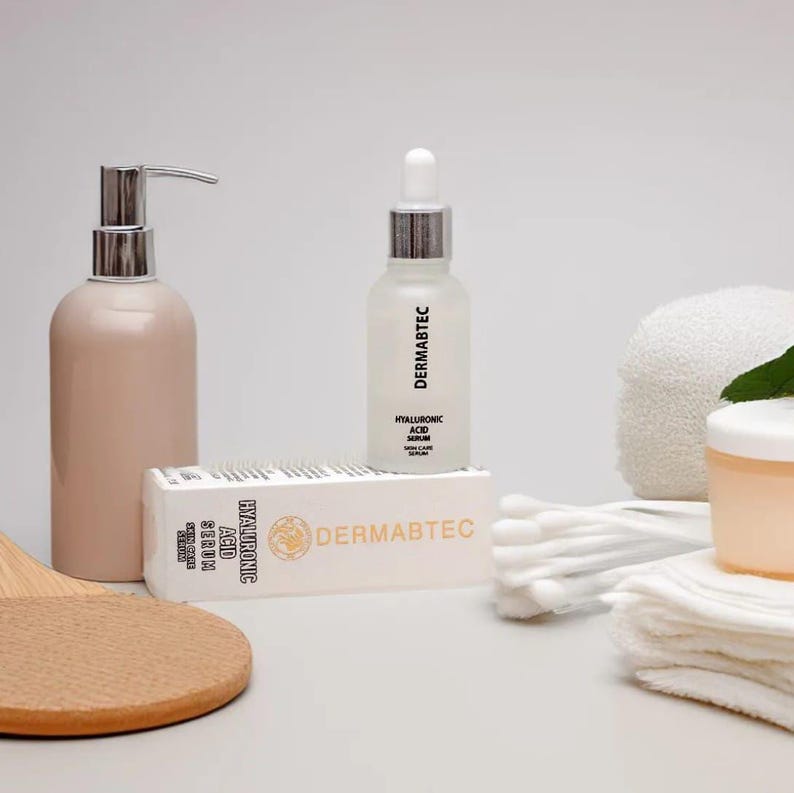 May include: A skincare product arrangement featuring a light pink pump bottle, a clear serum bottle with a dropper, and a box labeled "DERMABTEC HYALURONIC ACID SERUM". Additional items include cotton swabs, a small jar, and a white towel.