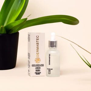 May include: A skincare product display featuring a frosted glass bottle with a dropper and a white box. The box and bottle both have the brand name "DERMABTEC" and the words "COLLAGEN-ANTI AGING SERUM SKIN CARE SERUM". A green plant is in the background.