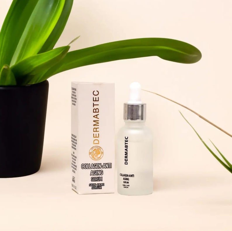 May include: A skincare product display featuring a frosted glass bottle with a dropper and a white box. The box and bottle both have the brand name "DERMABTEC" and the words "COLLAGEN-ANTI AGING SERUM SKIN CARE SERUM". A green plant is in the background.