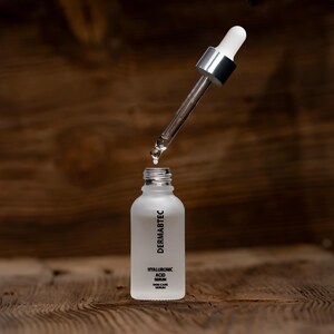 May include: A frosted glass bottle with a dropper dispensing a clear liquid. The bottle has the text "DERMABTEC" and "HYALURONIC ACID SERUM SKIN CARE SERUM". The dropper is silver and white, and the background is a wooden surface.