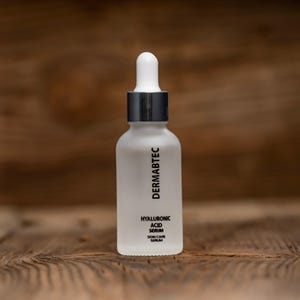 May include: A frosted white glass bottle with a silver collar and white dropper. The bottle has the text "DERMABTEC" printed vertically, and "HYALURONIC ACID SERUM SKIN CARE SERUM" printed below. The bottle is on a wooden surface.