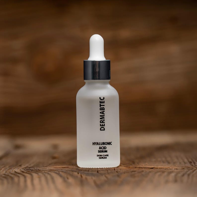 May include: A frosted white glass bottle with a silver collar and white dropper. The bottle has the text "DERMABTEC" printed vertically, and "HYALURONIC ACID SERUM SKIN CARE SERUM" printed below. The bottle is on a wooden surface.