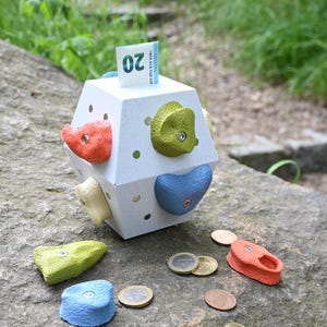 May include: A white, multi-faceted geometric money box featuring colourful, textured climbing holds attached to its sides. A twenty-euro note is inserted into the top slot, and several loose coins and additional climbing holds are scattered nearby.