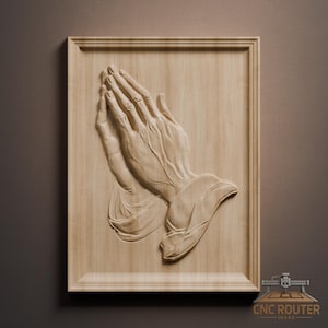 May include: A wooden wall art piece featuring a detailed carving of two hands clasped in prayer. The hands are intricately carved, showcasing the veins and contours. The artwork is framed in a light-colored wood, creating a three-dimensional effect.