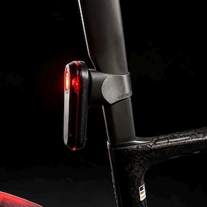 Pode incluir: Um quadro de bicicleta preto com textura de fibra de carbono e uma luz traseira LED vermelha montada no espigão do selim. O pneu traseiro é um modelo Continental Grand Prix 5000 preto. Um suporte de garrafa preto está montado no quadro.