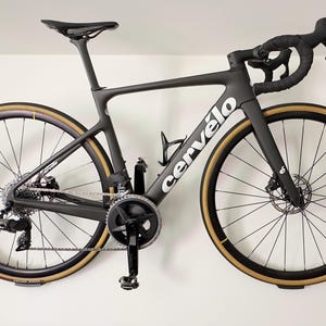 Peut inclure: Vélo de route Cervélo noir monté sur un mur blanc. Le vélo a des pneus à flancs beiges, un cadre noir et le logo Cervélo en blanc. Le guidon est noir, et il y a un porte-bidon.