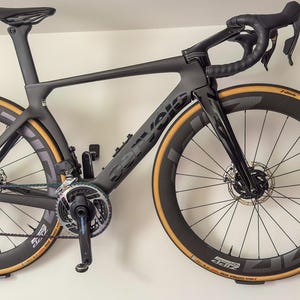 Peut inclure: Un vélo de route noir avec un cadre noir et des roues noires avec des flancs couleur sable. Le vélo est accroché à un mur.