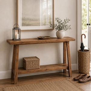 May include: A rectangular wooden console table with a lower shelf, featuring a dark metal lantern with a white candle, a shallow brown bowl, and a white ribbed vase with greenery on top, plus a woven storage box on the lower shelf.