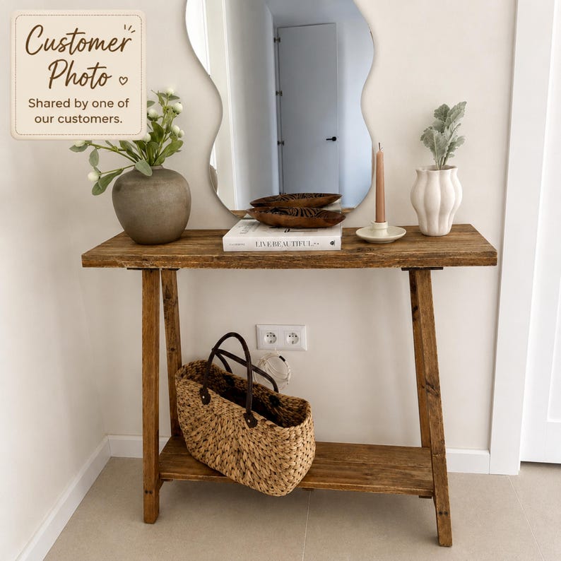 May include: A rustic wooden console table with a lower shelf, featuring a grey vase with white flowers, a book titled Live Beautiful, a brown wooden bowl, a beige taper candle in a holder, and a white fluted vase with greenery. A woven basket sits below.