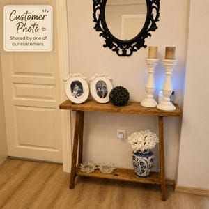 May include: A wooden console table with a lower shelf, featuring a natural wood grain finish. The table holds two white oval picture frames, a small green topiary ball, two white pillar candle holders, and a blue and white patterned vase.