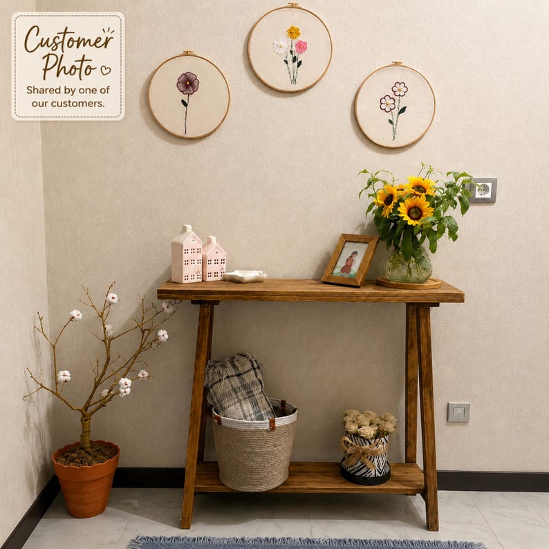 May include: A wooden console table with a lower shelf, holding a glass vase of sunflowers, a small framed picture, and two house-shaped ornaments. Below, a woven basket contains a plaid blanket and a decorative pot of white flowers.