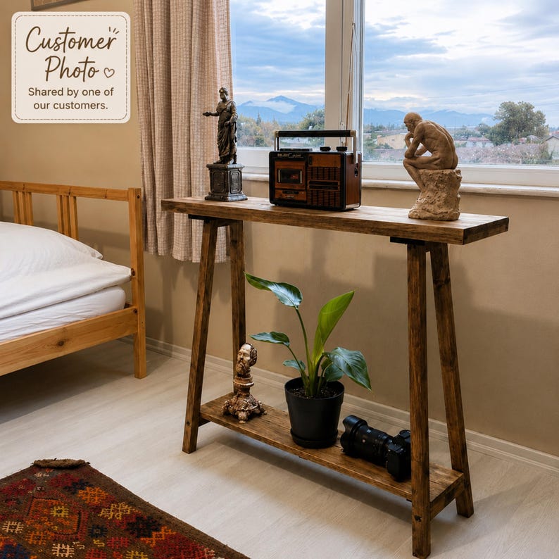May include: A rectangular wooden console table with an A-frame base and a lower shelf. The top surface holds a vintage-style radio, a small statue, and a sculpture of a seated figure. The lower shelf holds a potted green plant and a camera lens.