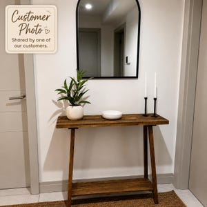May include: A rectangular wooden console table with a lower shelf, featuring a dark brown finish. On the table sits a green plant in a white ceramic pot, a white bowl, and two tall black candle holders with white taper candles. An arched mirror hangs above.