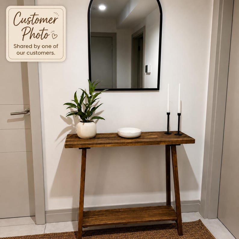 May include: A rectangular wooden console table with a lower shelf, featuring a dark brown finish. On the table sits a green plant in a white ceramic pot, a white bowl, and two tall black candle holders with white taper candles. An arched mirror hangs above.