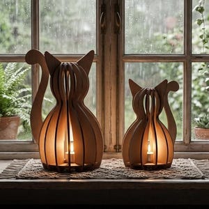 Op de afbeelding: Twee katvormige lampen gemaakt van verticale lichtgekleurde houten latten. Elke lamp heeft een hol centrum met een kleine gloeiende lichtbron. De katfiguren hebben puntige oren en gekrulde staarten en staan op een geweven mat.