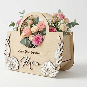 May include: A wooden flower basket with a handle, filled with pink and cream roses, eucalyptus, and baby's breath. The front of the basket has the words "Love You Forever Mom" and decorative floral cutouts.