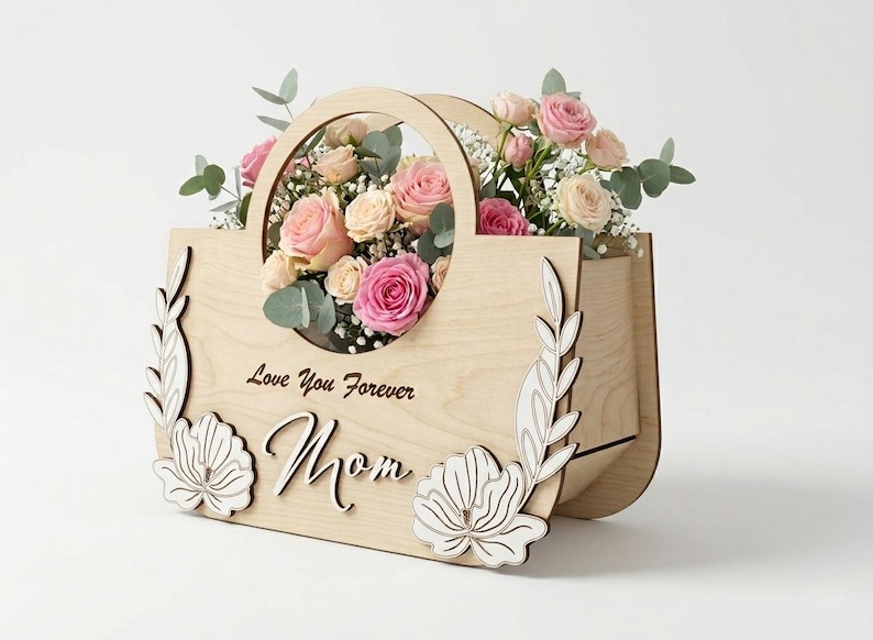 May include: A wooden flower basket with a handle, filled with pink and cream roses, eucalyptus, and baby's breath. The front of the basket has the words "Love You Forever Mom" and decorative floral cutouts.