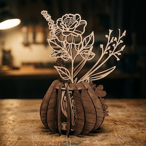 Può includere: Composizione floreale in legno con una rosa, foglie e altri elementi botanici. La composizione è inserita in una struttura a forma di vaso con finitura marrone scuro. Il design è intricato, con dettagli ritagliati. La parola "MOM" è visibile.