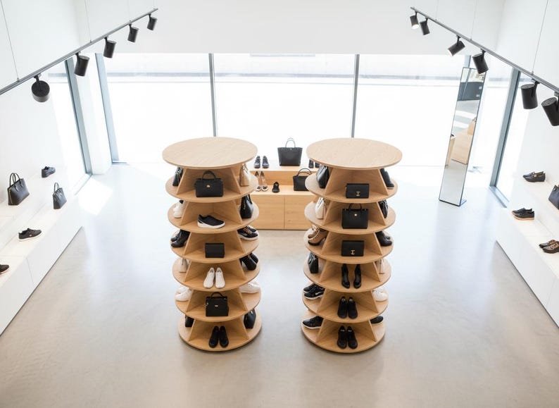 K&ouml;nnte beinhalten: Moderne Ladenauslage mit zwei kreisf&ouml;rmigen Holzregalen, die Schuhe und Handtaschen pr&auml;sentieren. Die Regale sind spiralf&ouml;rmig angeordnet, wobei jede Ebene verschiedene Stile zeigt. Der Laden hat ein minimalistisches Design mit heller Beleuchtung.