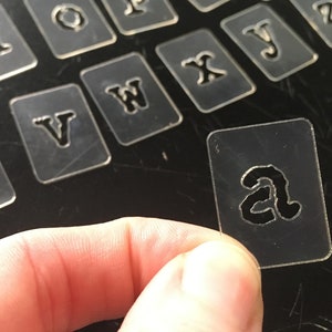 May include: A set of clear plastic stencils with different letters of the alphabet. The stencils are arranged in a grid pattern on a black surface. The letter "a" is being held by a finger.