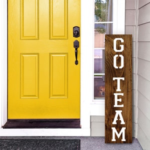 K&ouml;nnte beinhalten: Ein Holzschild mit den Worten "GO TEAM" in wei&szlig;en Buchstaben. Das Schild lehnt an einer wei&szlig;en T&uuml;r mit einer gelben T&uuml;r dahinter.
