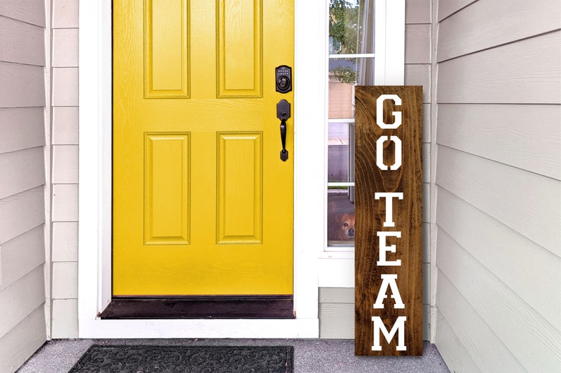 K&ouml;nnte beinhalten: Ein Holzschild mit den Worten "GO TEAM" in wei&szlig;en Buchstaben. Das Schild lehnt an einer wei&szlig;en T&uuml;r mit einer gelben T&uuml;r dahinter.