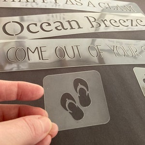 May include: A set of four clear plastic stencils with various beach-themed phrases and images. The stencils include the phrases "Ocean Breeze", "Come Out of Your Shell", and an image of flip flops.