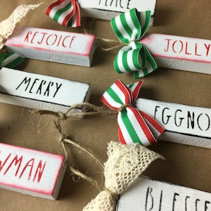 May include: Set of eight wooden Christmas ornaments with red and green ribbon bows. Each ornament has a different Christmas-themed word written on it: "Peace", "Rejoice", "Jolly", "Merry", "Eggnog", "Snowman", "Blessed", and "Believe".