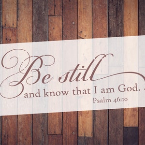 May include: A stencil with the words "Be still and know that I am God. Psalm 46:10" in a brown script font. The stencil is on a white background, set against a backdrop of wooden planks.