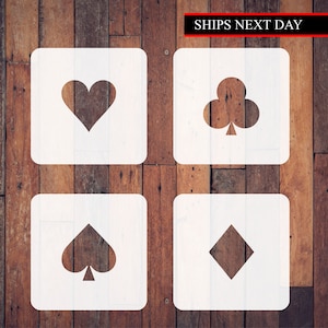 May include: Four white square stencils with black card suit symbols: heart, club, spade, and diamond. The stencils are on a wood background.