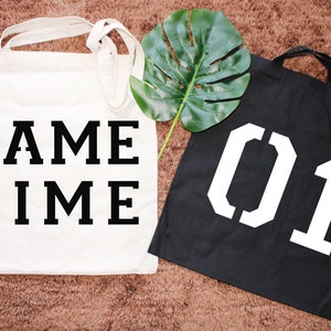 K&ouml;nnte beinhalten: Zwei wei&szlig;e Canvas-Tragetaschen mit schwarzem Text. Eine Tasche sagt "GAME TIME" und die andere sagt "01".